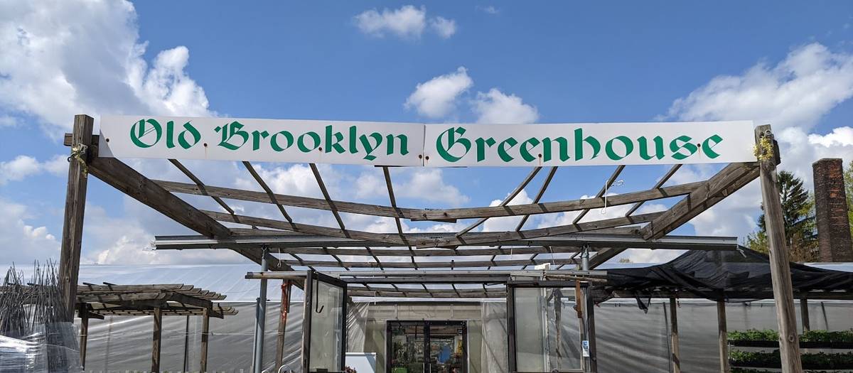 The painted green lettering of the Old Brooklyn Greenhouse sign over a wooden pergola at the front entrance, under a blue sky.